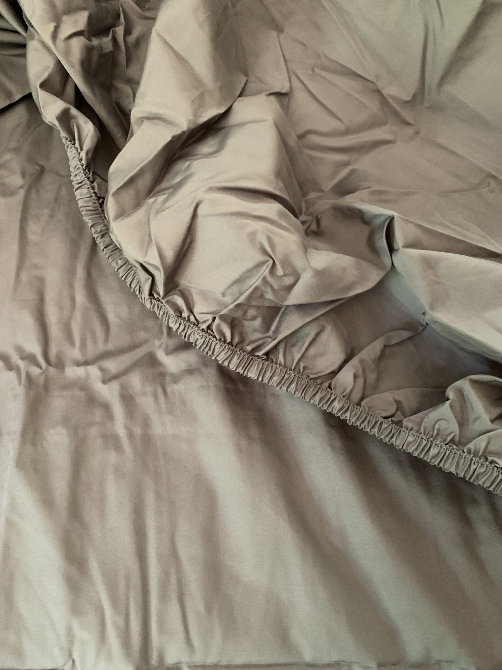 A close-up view of a beige fitted bed sheet with elastic edges, slightly rumpled, lying on a matching flat sheet.