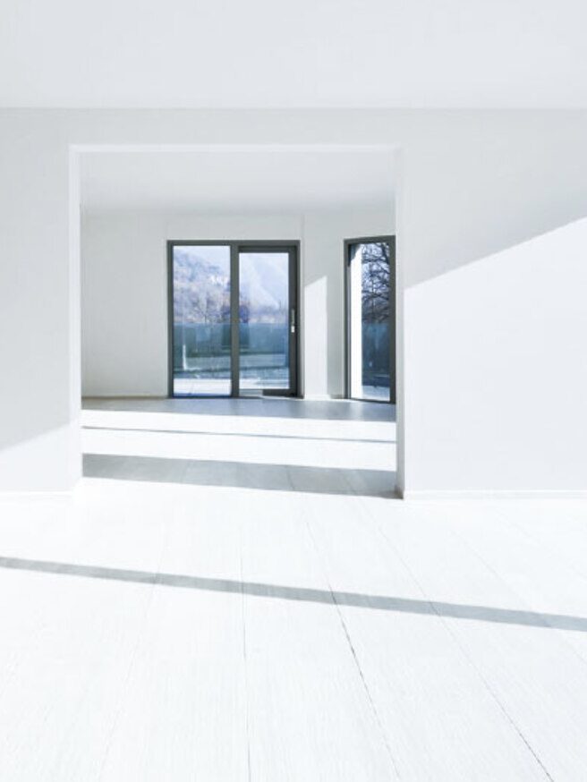 A bright, empty room with white walls and large windows allowing sunlight to stream in. The room features light-colored floors and an open doorway leading to another similarly lit area.