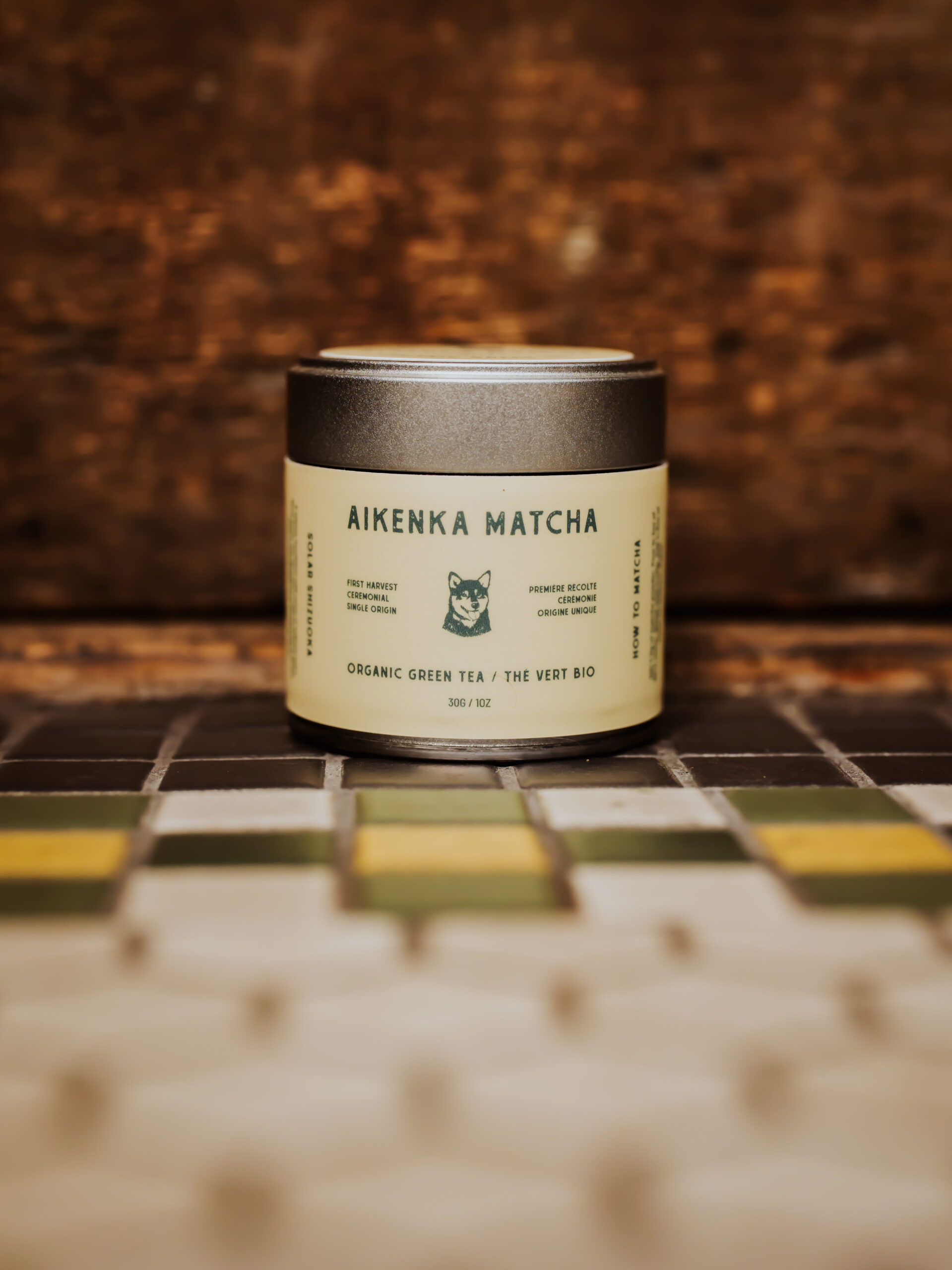 A container labeled "Aikenka Matcha Organic Green Tea" sits on a tiled surface with a wooden backdrop.