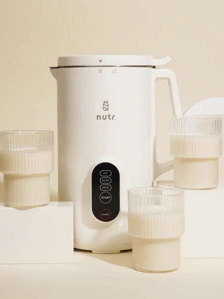 A white Nutr. appliance accompanied by three glasses filled with a creamy liquid, arranged on white blocks against a neutral background.