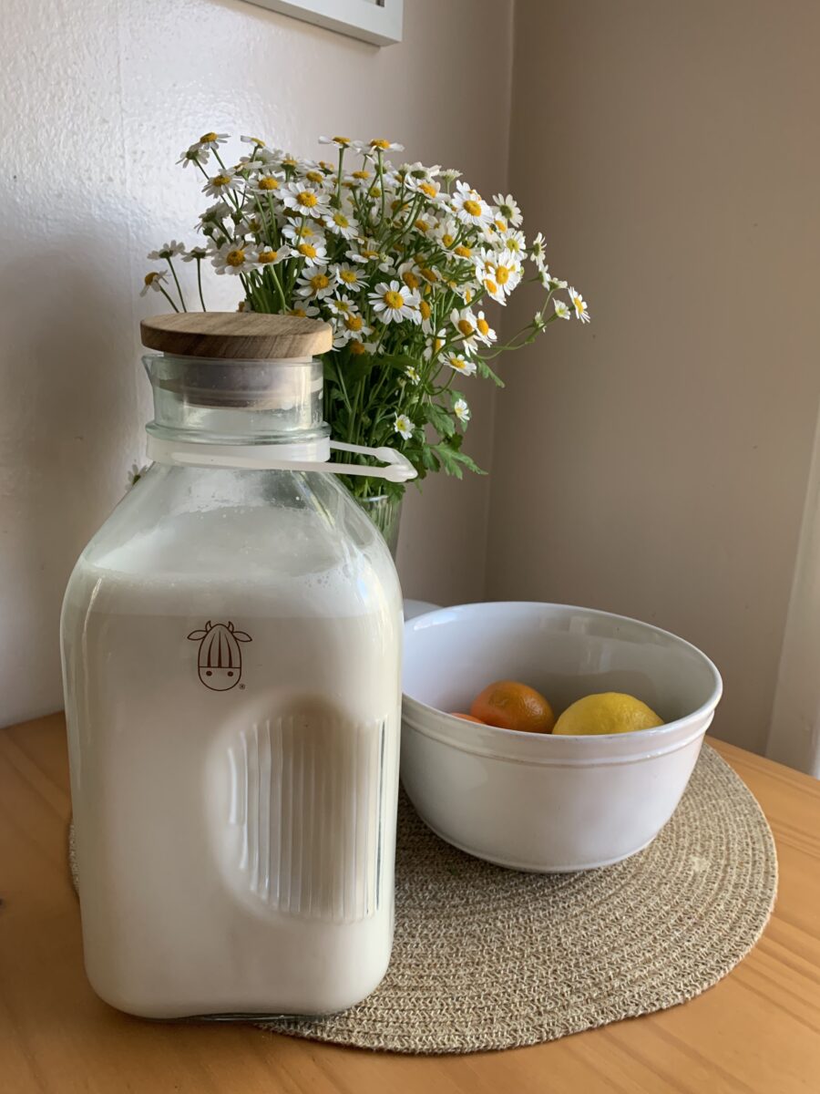 A glass jar of milk with a wooden lid is placed on a table next to a bowl containing lemons and an orange, with a vase of white flowers in the background.