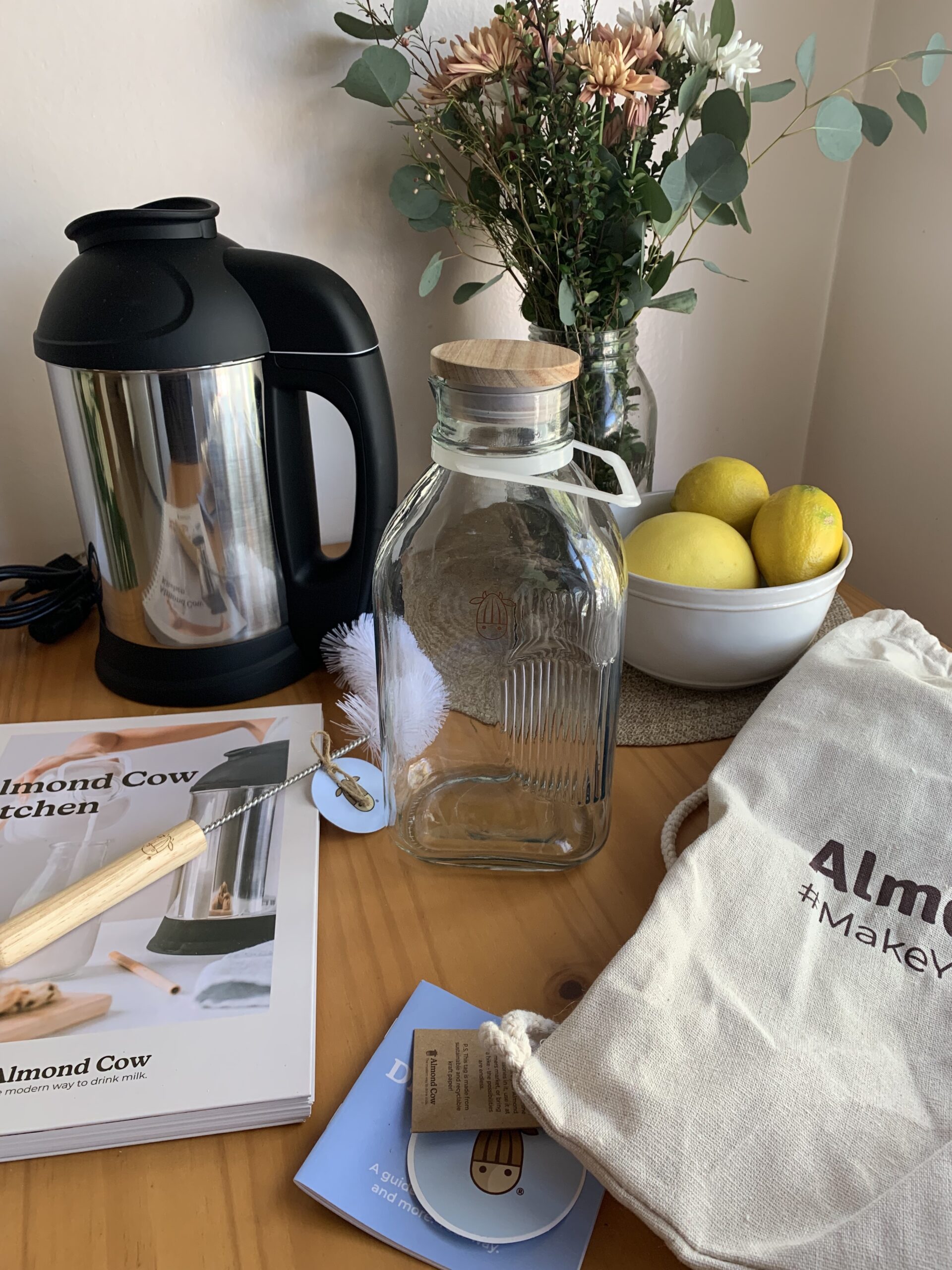 A countertop scene with a glass bottle, a black kitchen appliance, a bowl of lemons, a knife, a cleaning brush, a reusable bag, and various papers and books. Flowers in a vase are in the background.