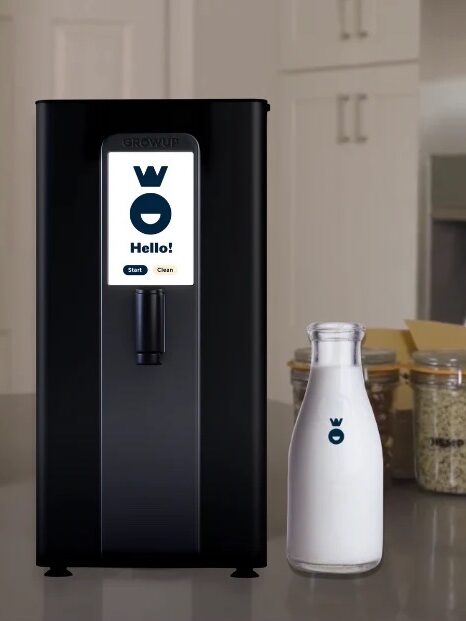 A black beverage dispenser with a digital "Hello!" display stands on a kitchen counter with a glass bottle of milk and jars of various food items in the background.