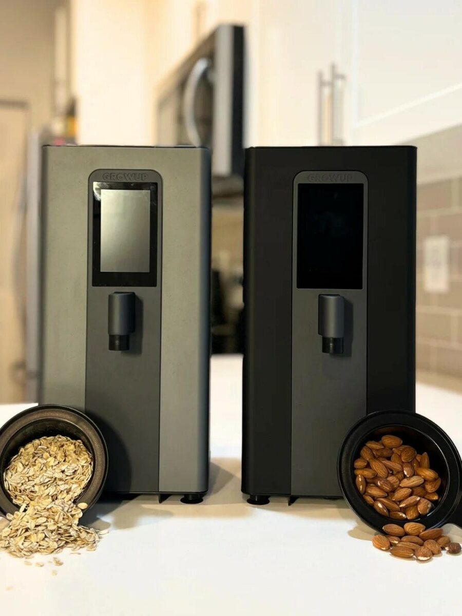 Two sleek countertop machines stand side by side in a kitchen, with a cup of oats in front of one and a cup of almonds in front of the other.