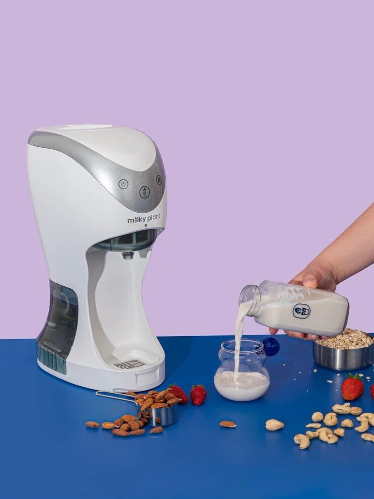 A person pours plant-based milk from a bottle into a glass with a countertop plant milk maker and assorted nuts and grains nearby.