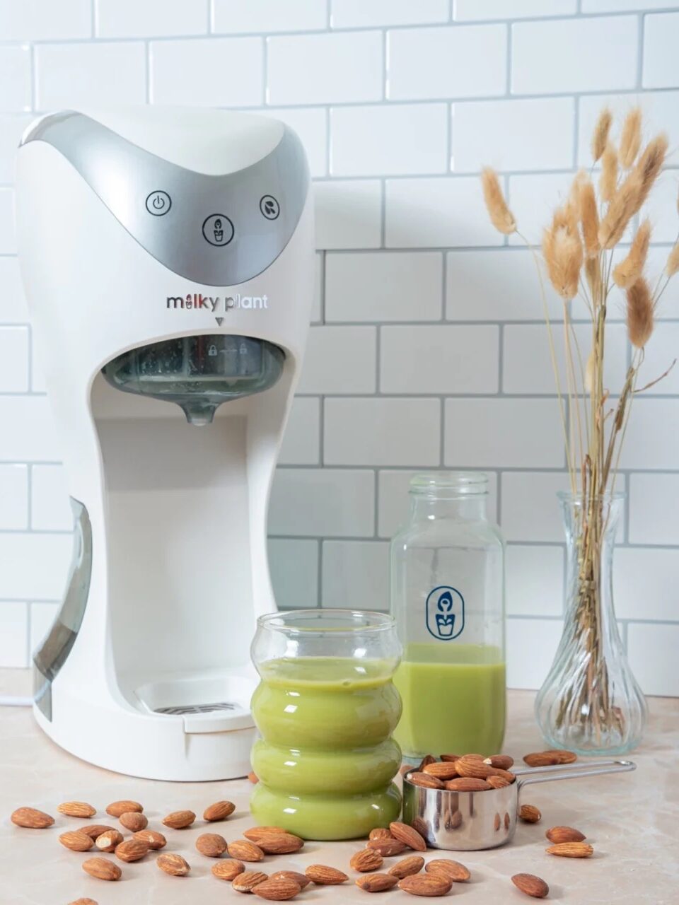 A white machine labeled 'Milky Plant' on a countertop next to a jar and a glass of green liquid, surrounded by almonds. A vase with dried grasses is in the background against a tiled wall.