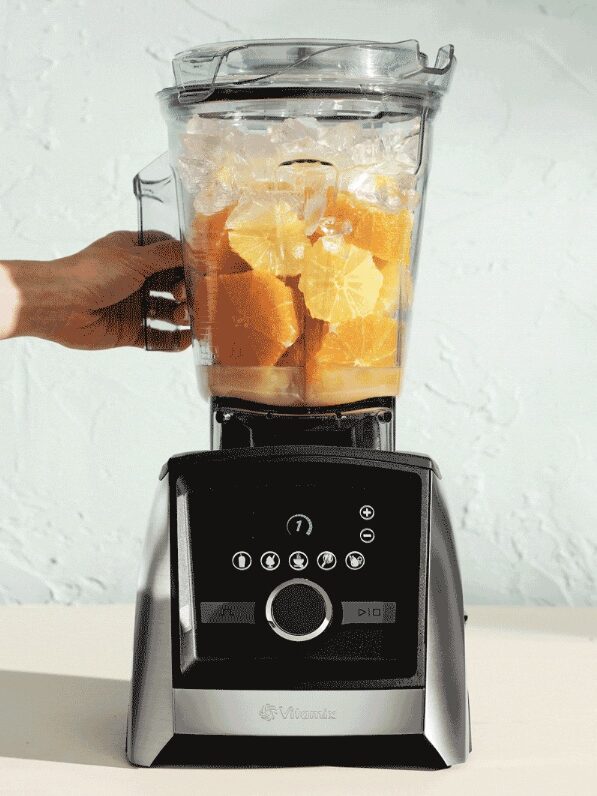 A hand places a blender jar filled with ice and orange slices onto a black and silver blender base against a light background.