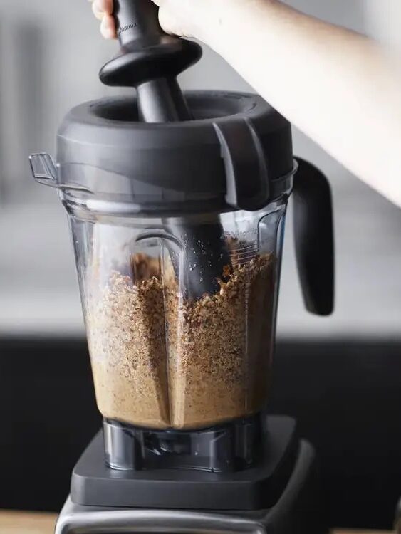 A person uses a pusher tool with a black blender to blend a brown mixture in a clear container placed on a kitchen countertop.