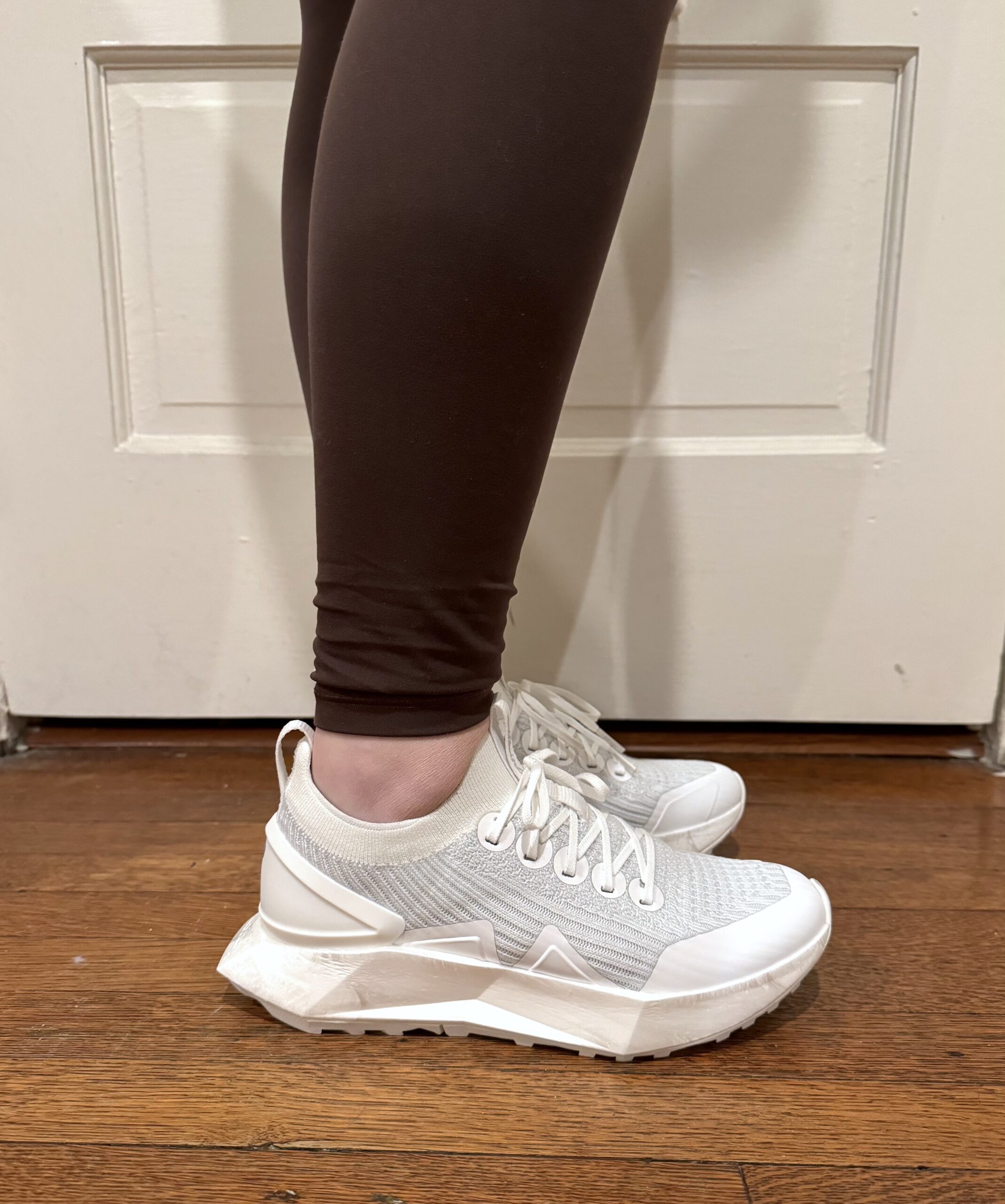 Person wearing white sneakers with thick soles and brown leggings stands on a wooden floor by a white door.