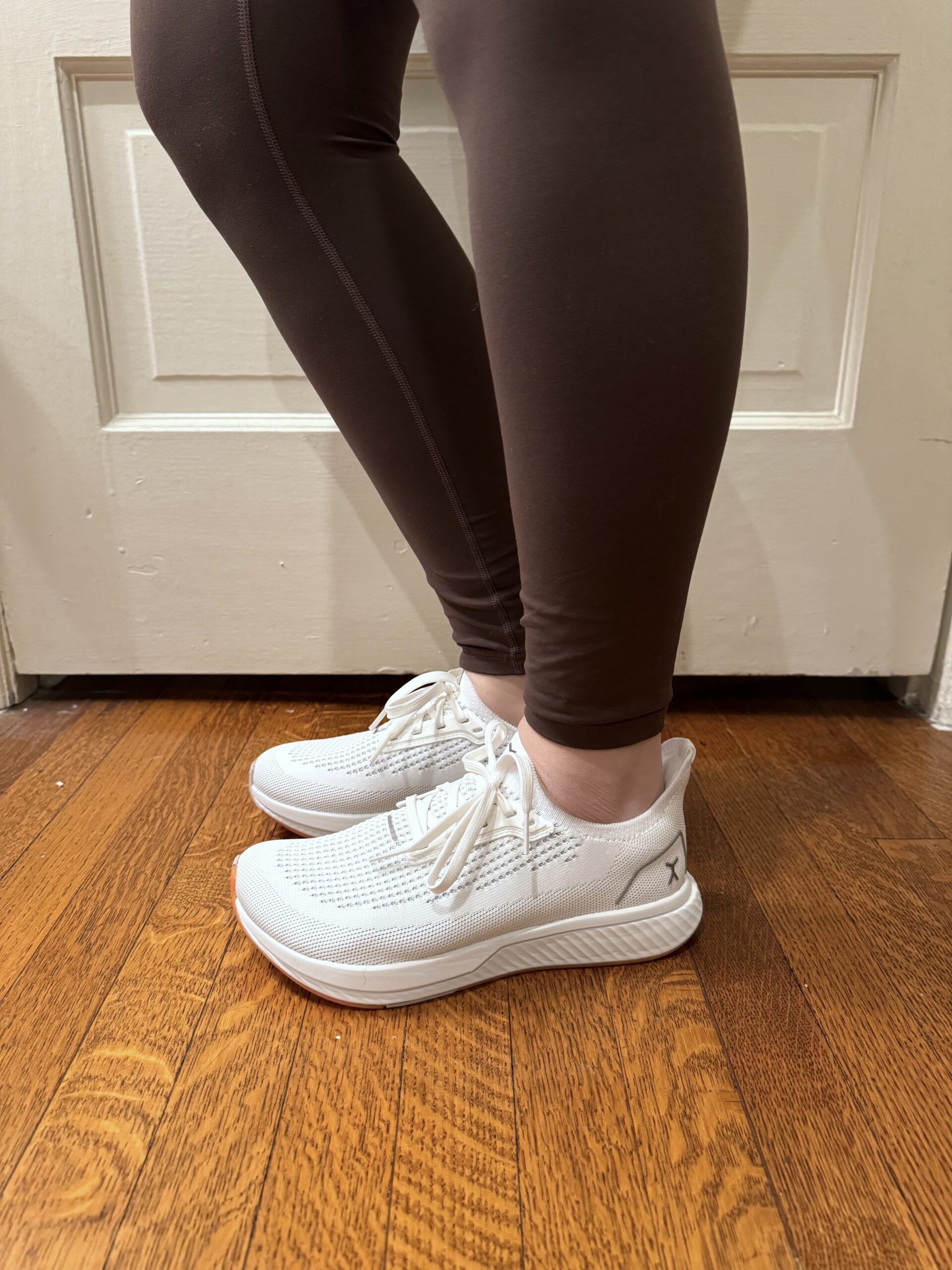 Person wearing white sneakers and black leggings stands on wooden floor in front of a white door.