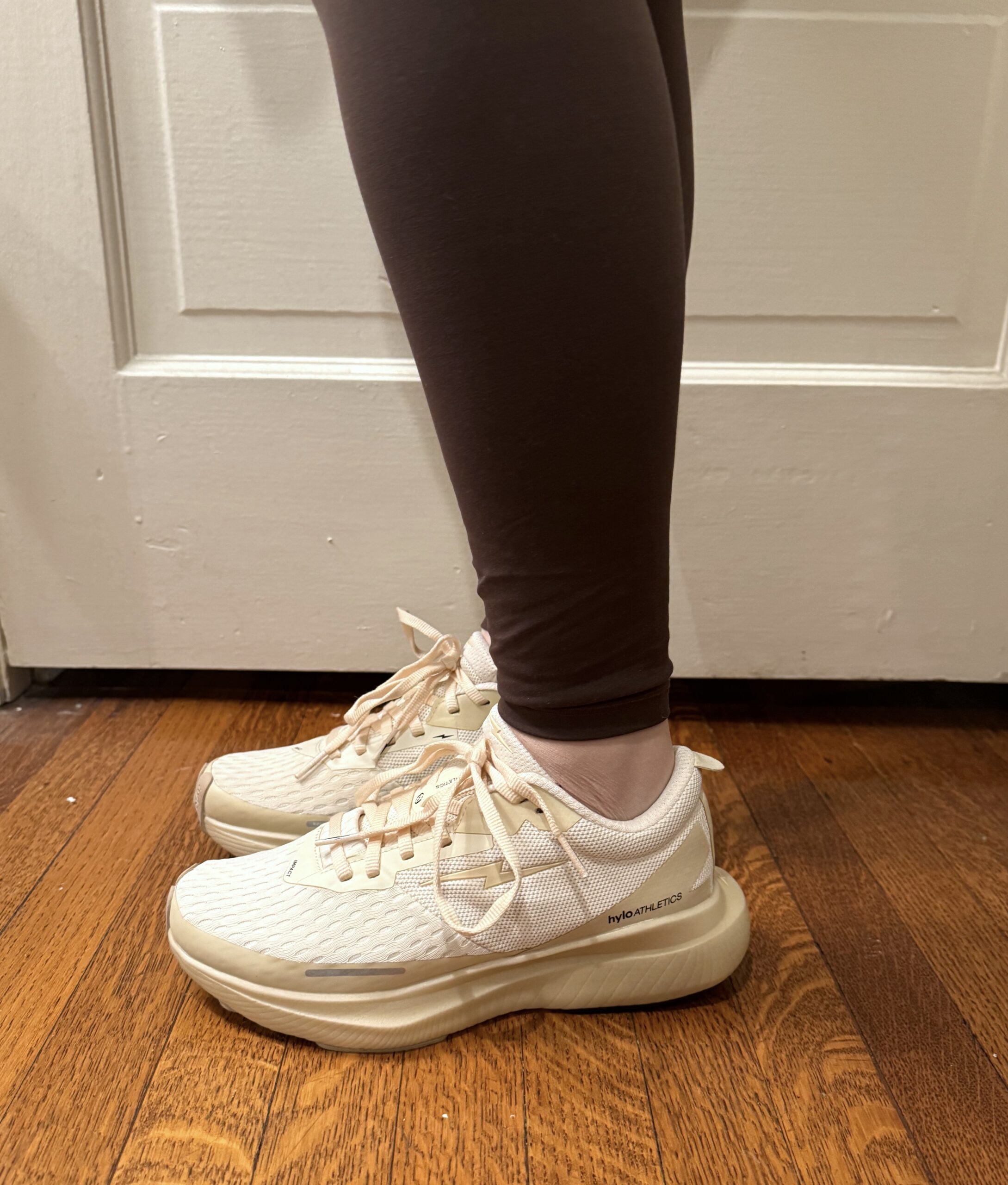 Person wearing cream-colored athletic shoes and dark leggings standing on a wooden floor.