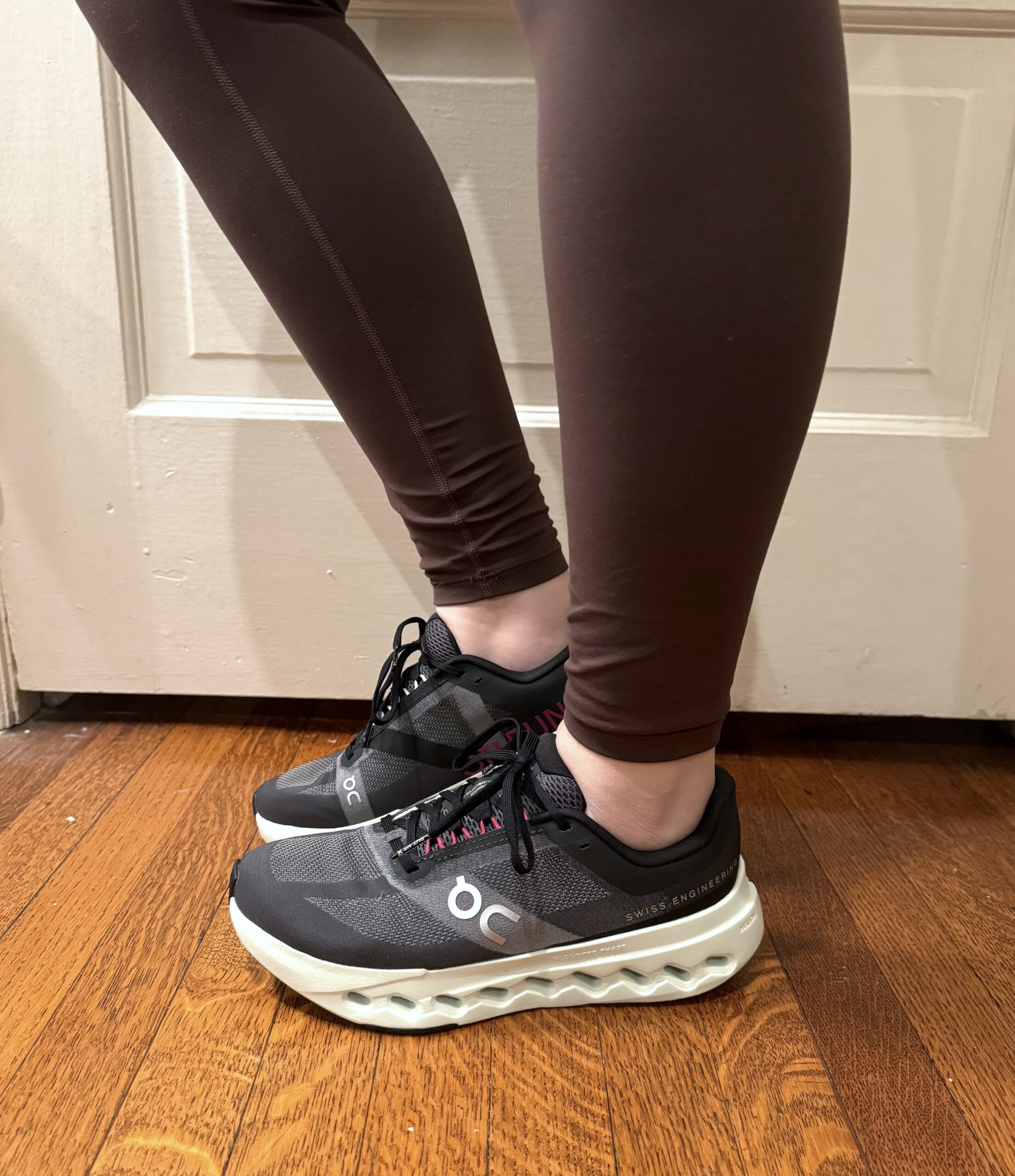 Person wearing black and gray athletic shoes with white soles, standing on a wooden floor in front of a white door.