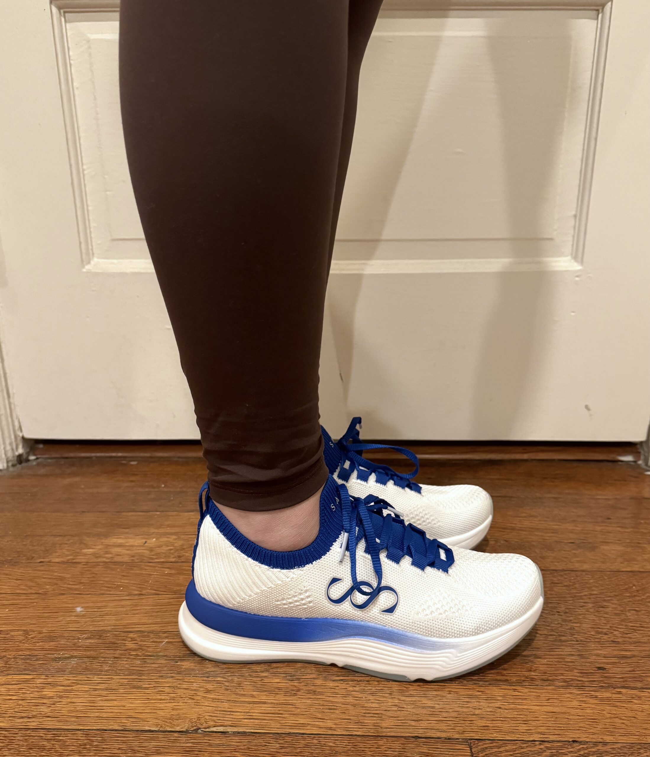 Person wearing white and blue athletic shoes and brown leggings standing on a wooden floor near a white door.