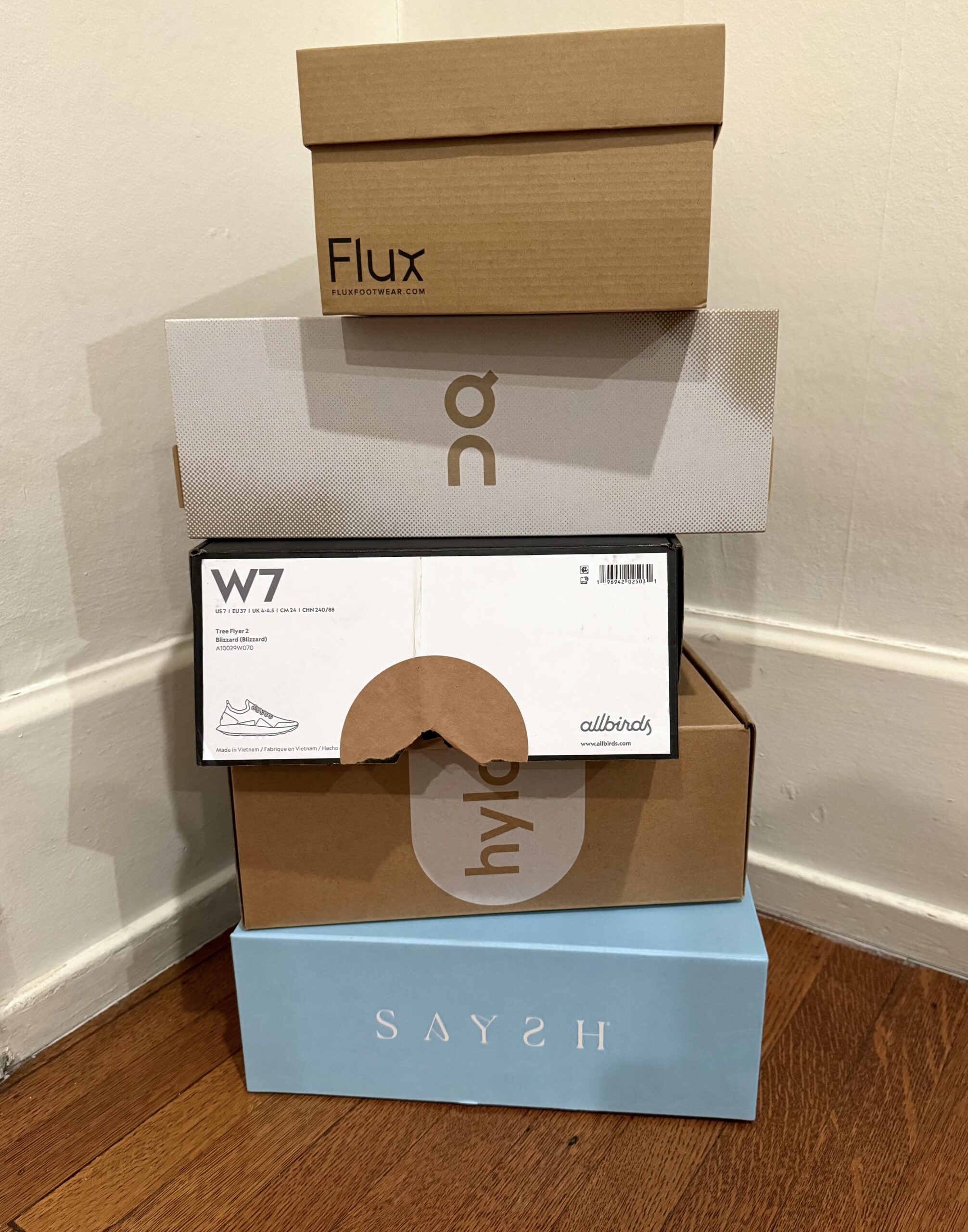 A stack of four shoe boxes from different brands, including Flux, Allbirds, and Saysh, placed on a wooden floor.