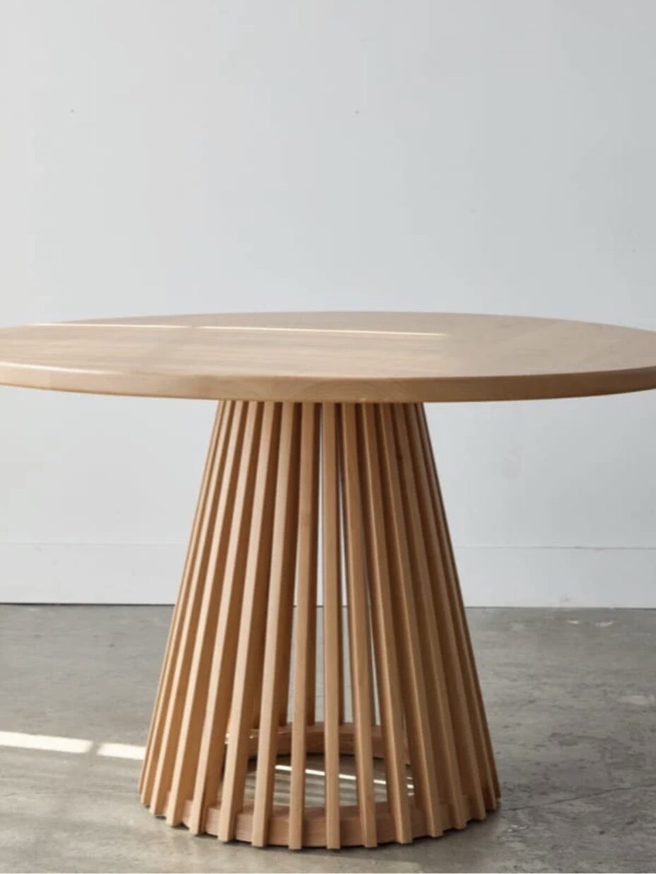 A round wooden table with a flat top and a base made of vertical slats arranged in a circular pattern, set against a plain white wall and a concrete floor.