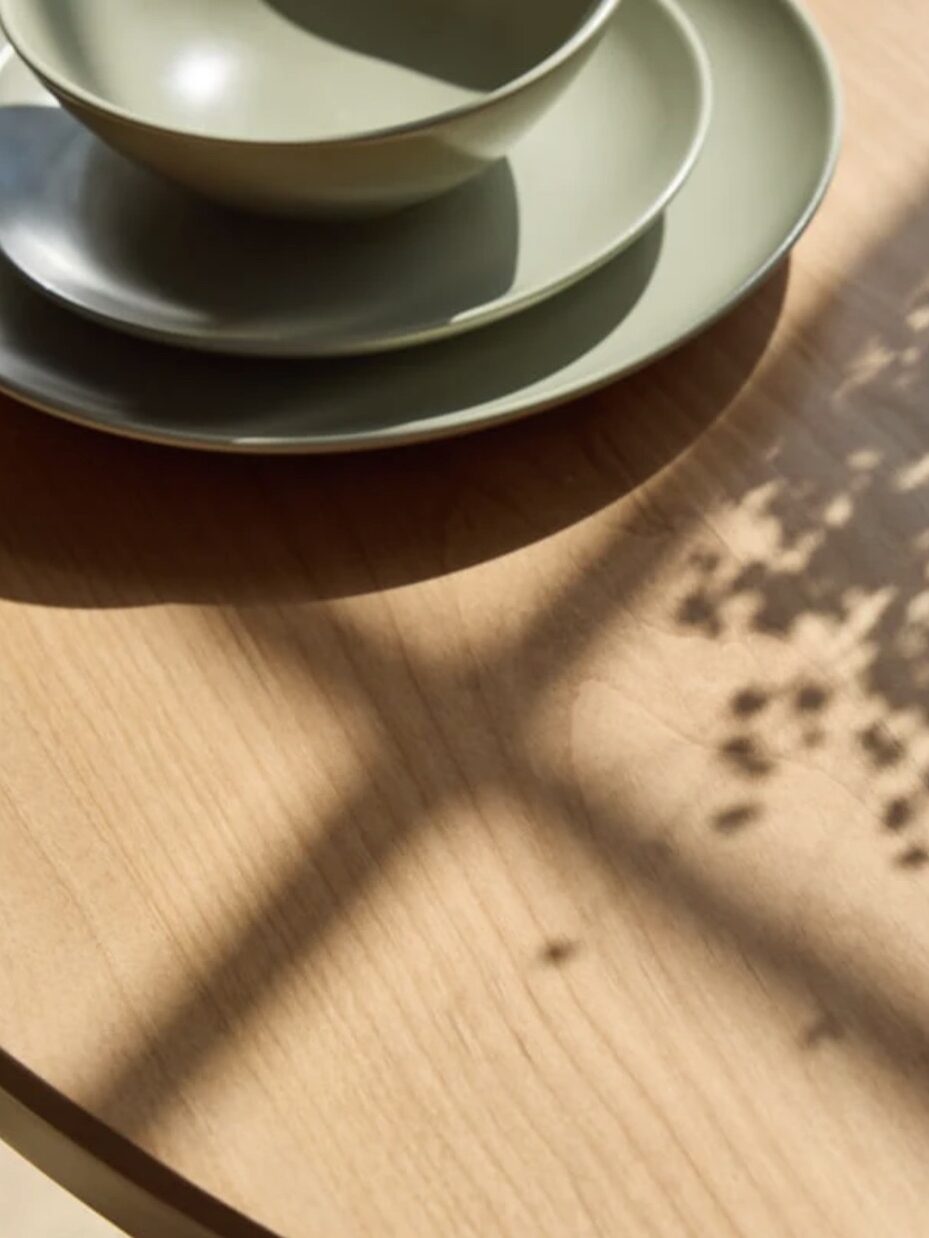 A wooden table with a stack of green ceramic plates and bowls, illuminated by sunlight and shadows from a window.