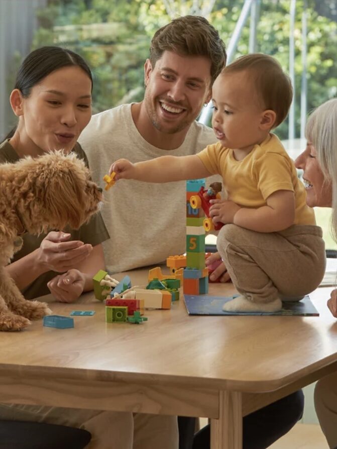 A baby sits on a table playing with toy blocks, surrounded by four adults and a dog. One adult holds the dog while two others look on, and the baby extends a toy towards the dog.