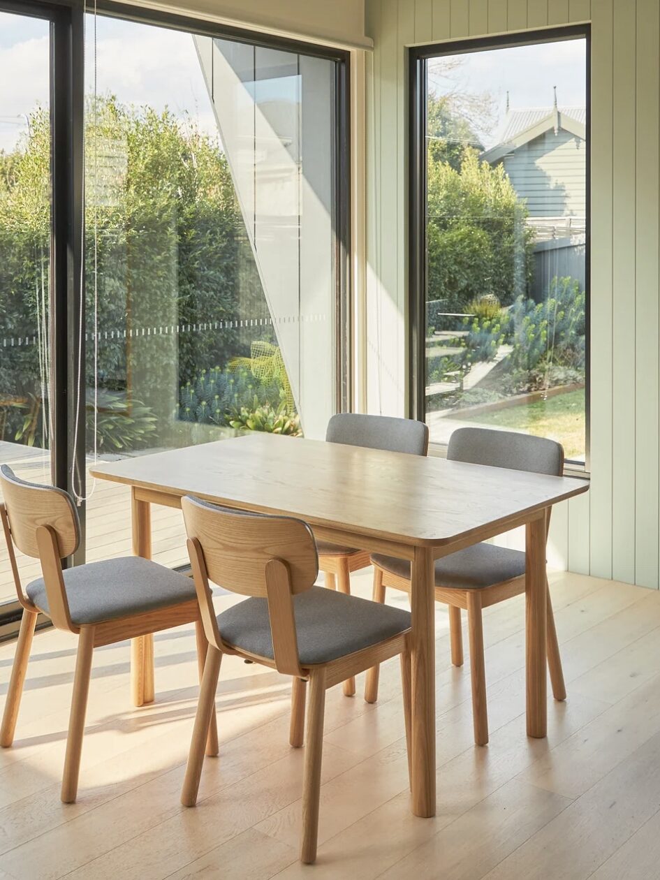 A wooden dining table with four matching chairs is situated in a room with large windows overlooking a garden.