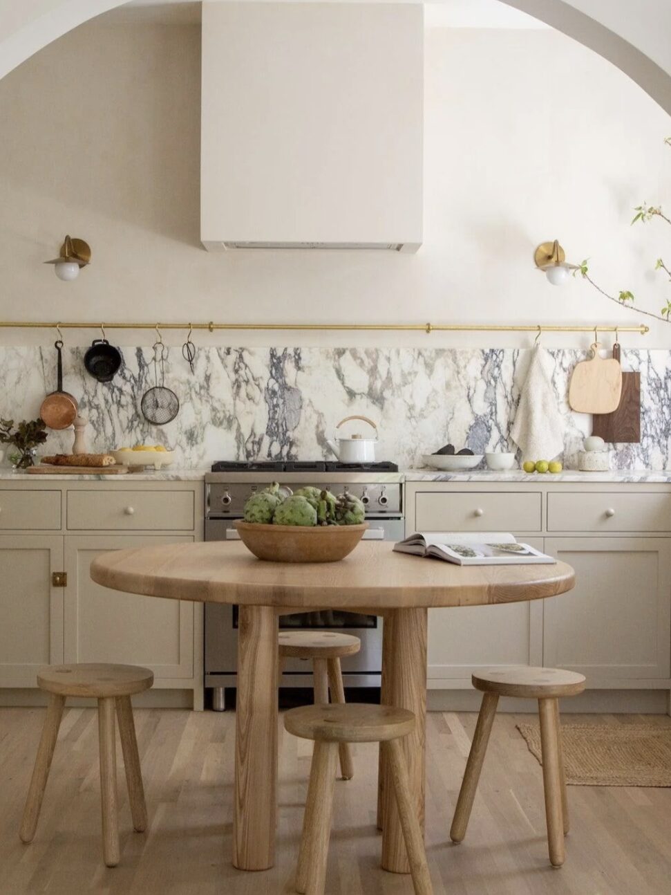 A modern kitchen with a marble backsplash, light wood cabinets, and stainless steel stove. A round wooden table with stools occupies the center, and plants add a touch of green to the setting.