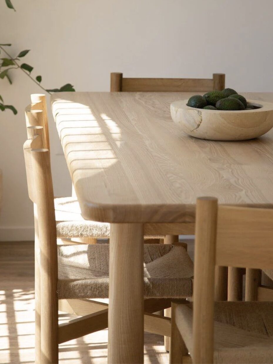 A wooden dining table with chairs is illuminated by sunlight. A wooden bowl filled with green avocados sits on the table. A leafy plant is partially visible in the background.