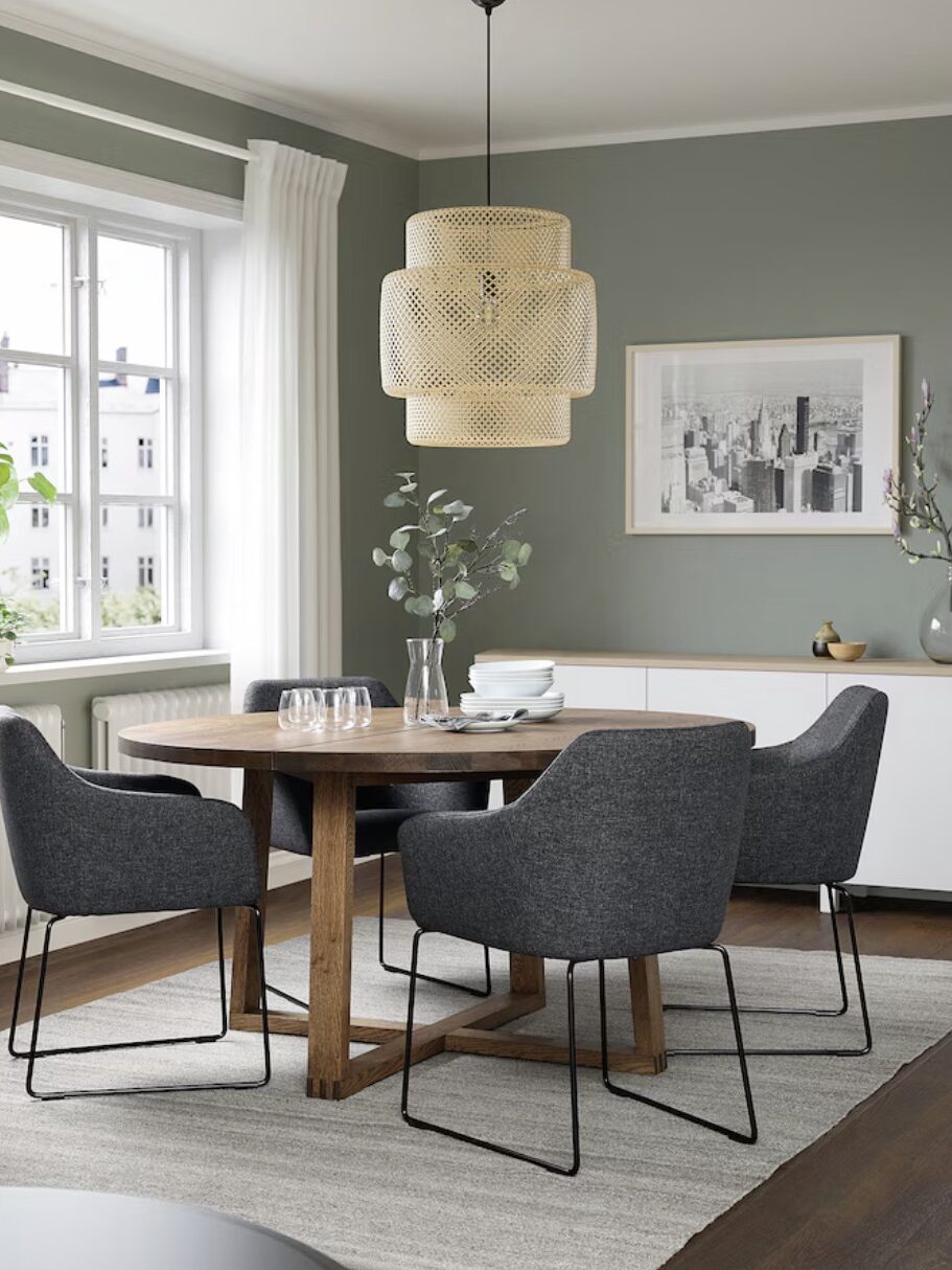 A modern dining room with a round wooden table, four gray upholstered chairs, a woven pendant light, potted plants, and a cityscape photo on the wall. Light green walls, a large window, and a sideboard are present.