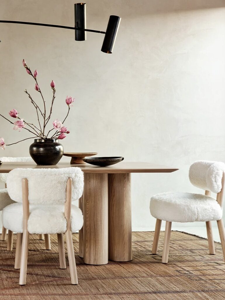 A dining area featuring a wooden table, four white, plush chairs, a vase with pink flowers, a wooden bowl, and a black bowl on a textured rug under a minimalist lighting fixture.