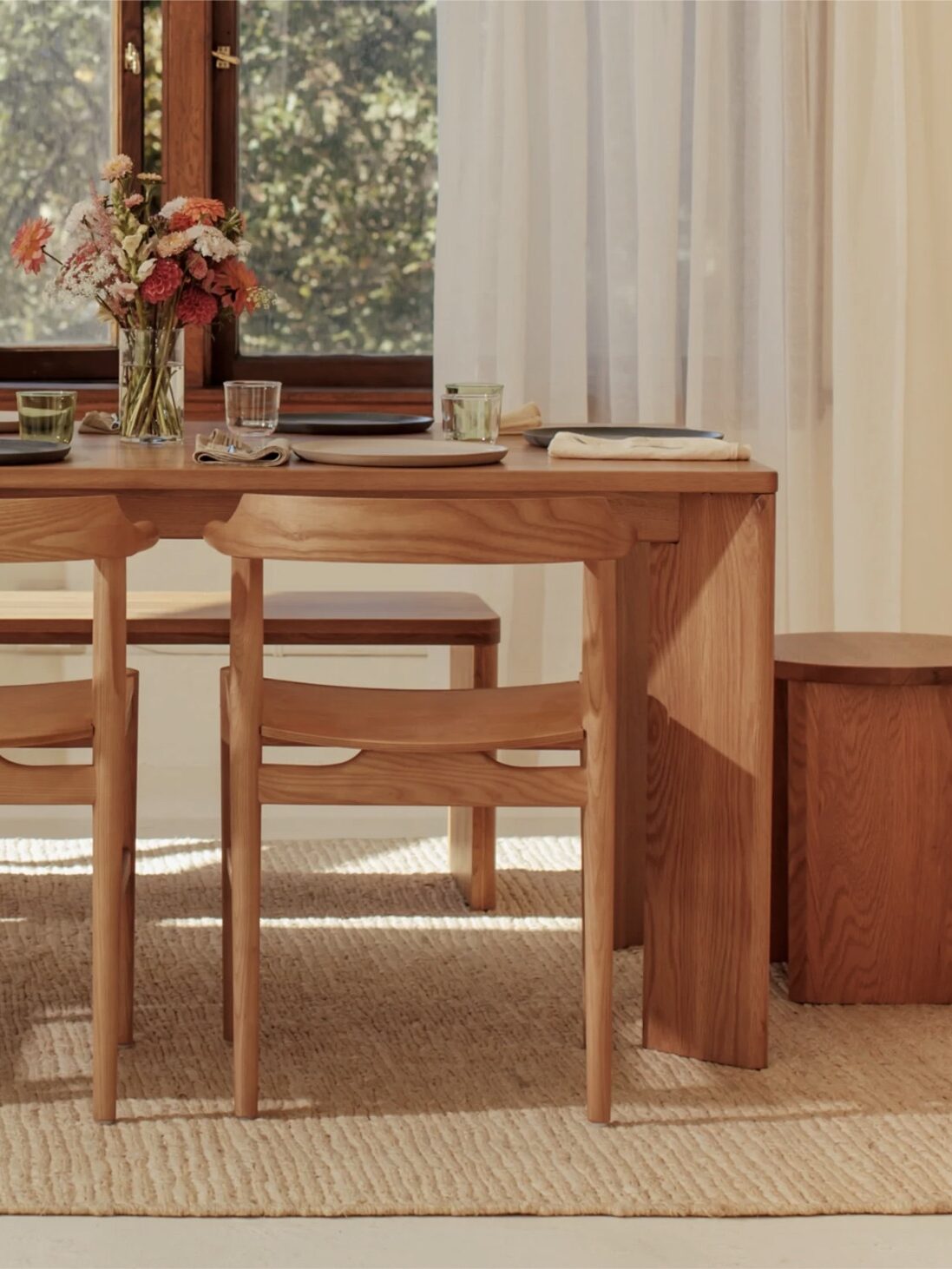 A wooden dining table with matching chairs is set with plates, glasses, and floral centerpieces. Sunlight filters through a window with sheer curtains. A woven rug covers the floor.