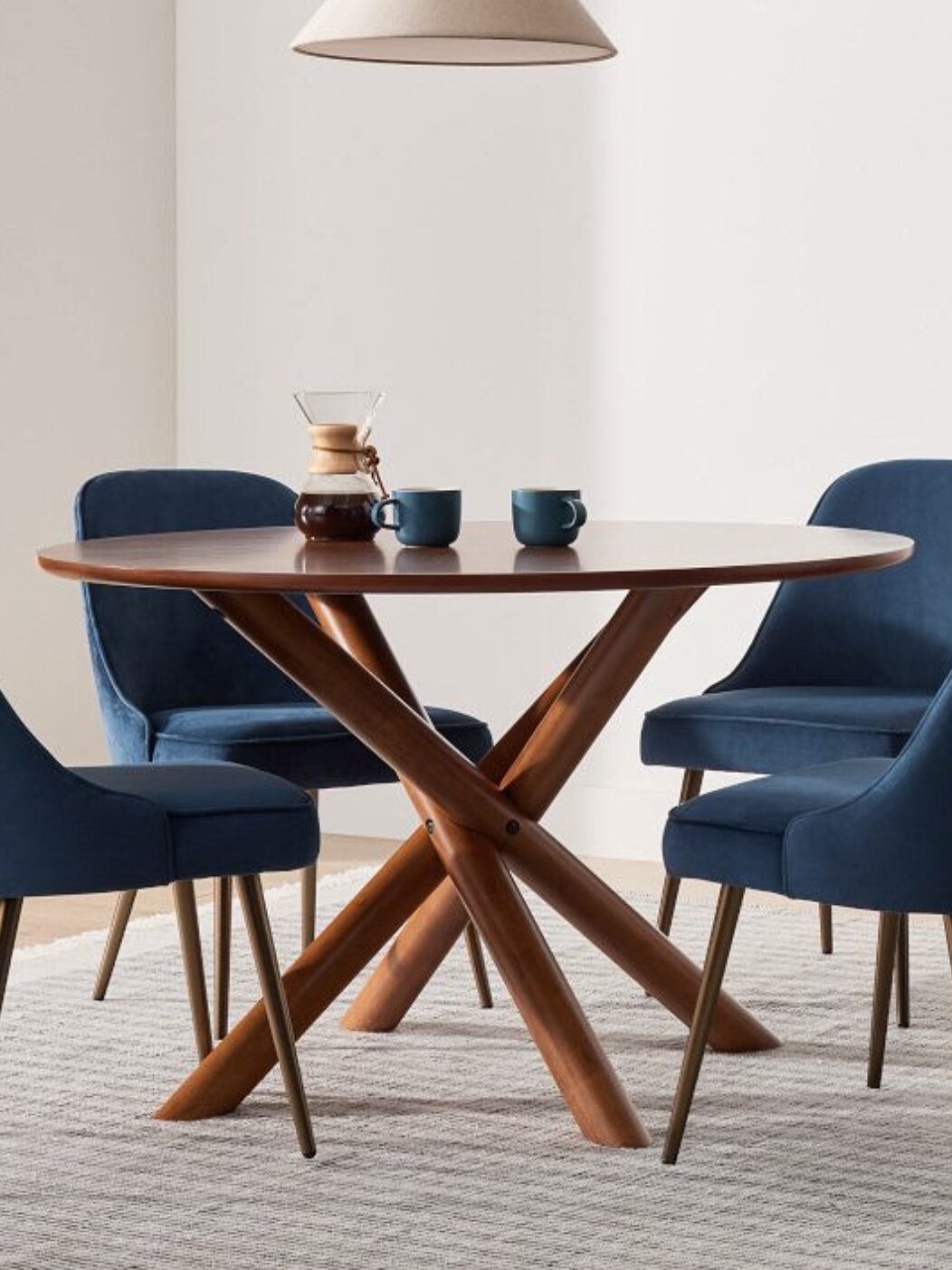 A round wooden table with four blue upholstered chairs, a coffee maker, and two blue mugs on top, set on a light-colored rug with a hanging light fixture above.