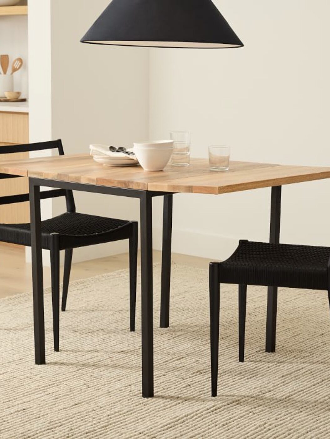 A small, rectangular dining table with a light wood top and black metal legs, accompanied by two black chairs, is set with a bowl, glass, and cup. A large, black, cone-shaped pendant light hangs above.