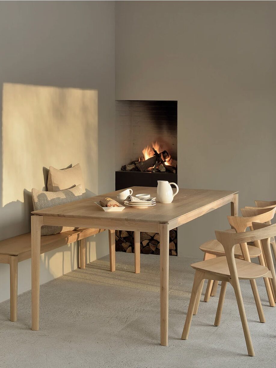 A minimalist dining area features a wooden table with four chairs, a beige bench with cushions, and a lit fireplace in the background.