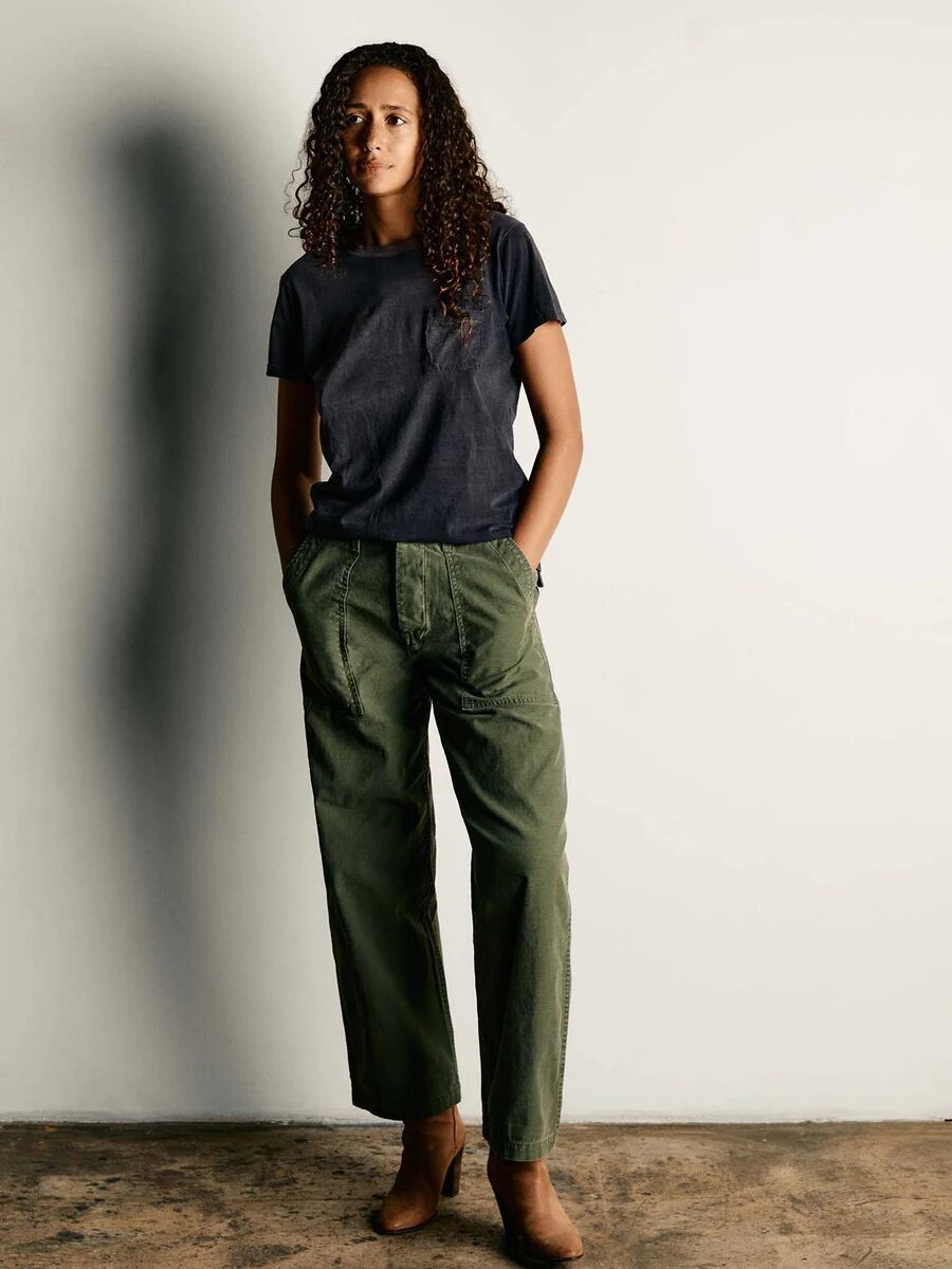 A person with curly hair stands against a plain backdrop, wearing a dark t-shirt, olive green pants, and brown shoes.