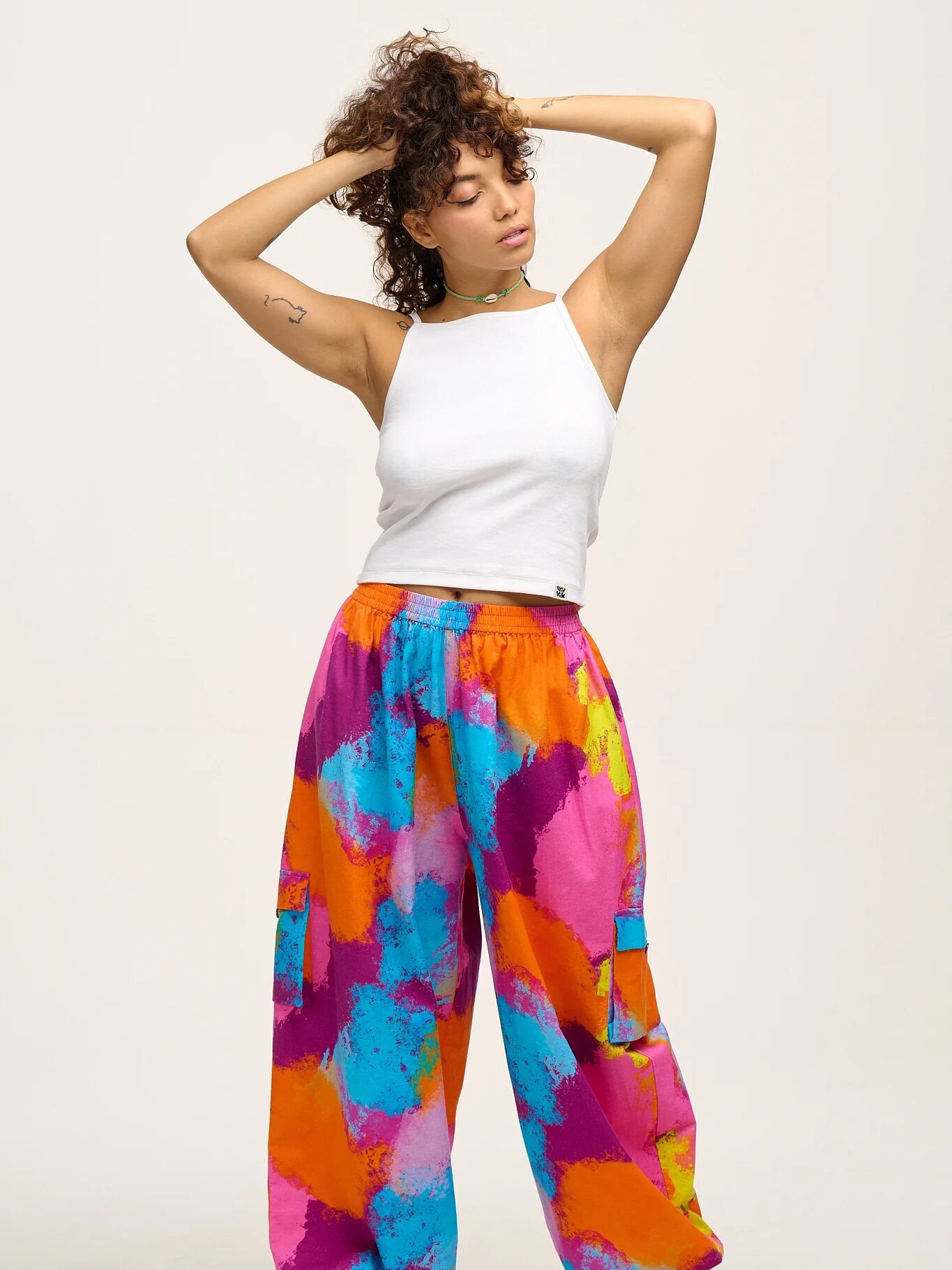 Person in a white sleeveless top and colorful tie-dye pants stands with hands behind their head against a plain background. They are wearing brown shoes and have curly hair.