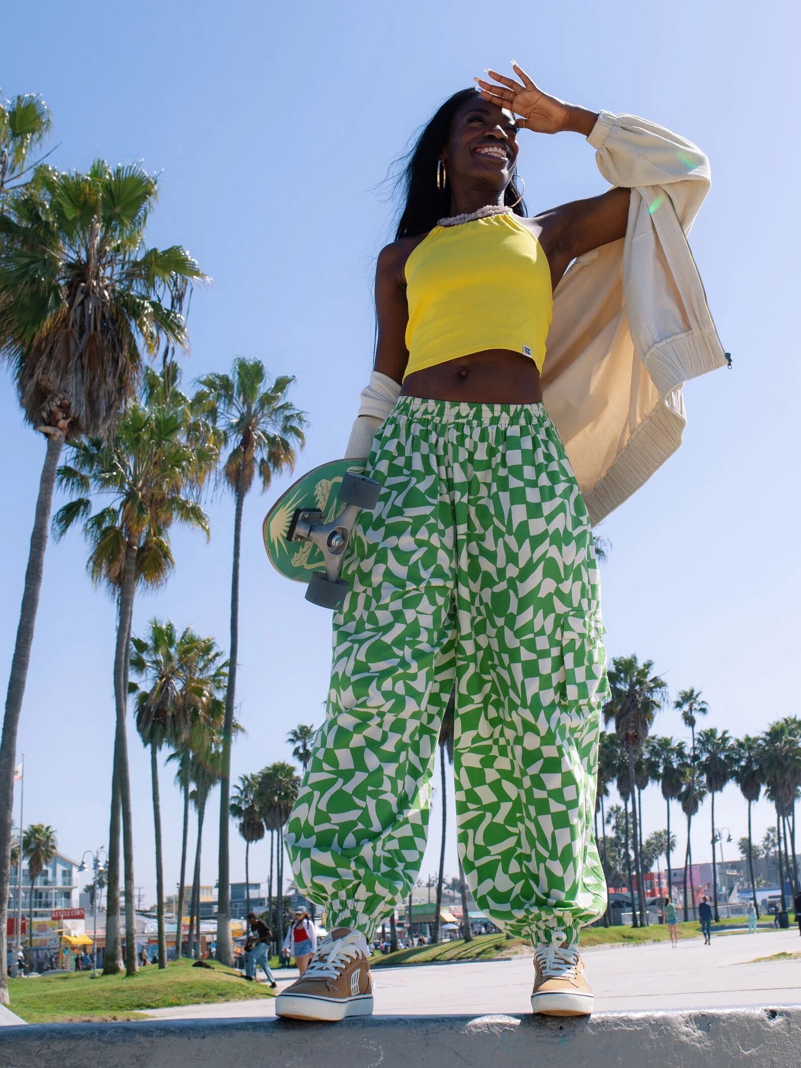 A person wearing a yellow top, patterned green and white pants, and tan sneakers stands outdoors, holding a skateboard and shielding their eyes from the sun, with palm trees in the background.