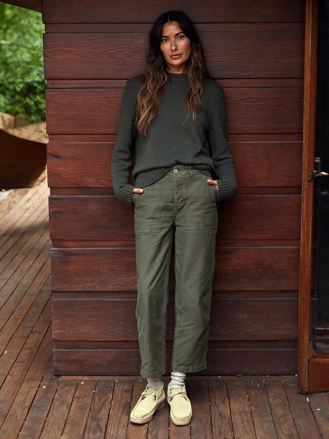 A woman with long hair stands on a wooden deck, leaning against a red wooden wall, with her hands in her pockets. She is wearing a dark green sweater, green pants, striped socks, and beige shoes.