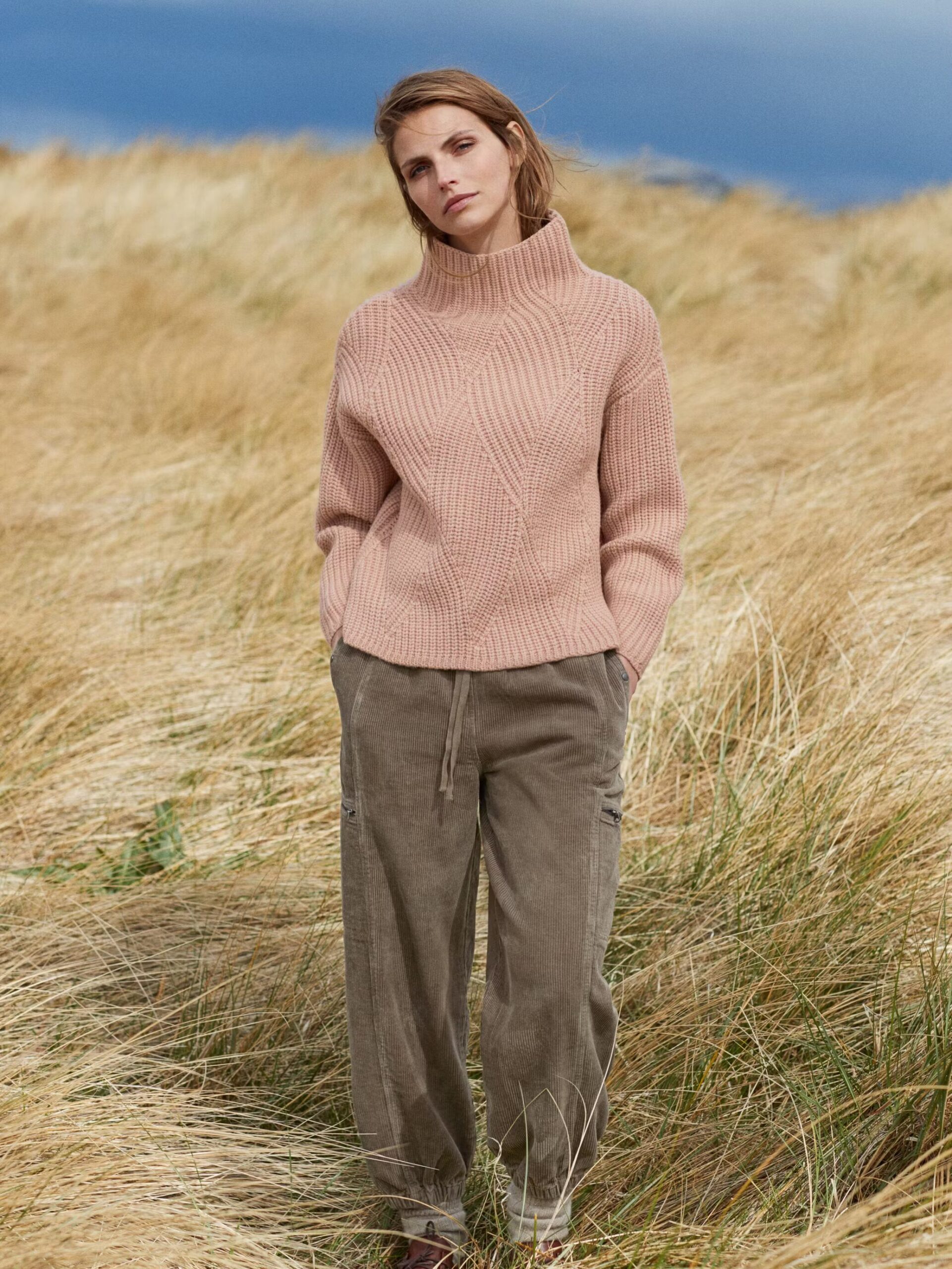 A person wearing a light brown turtleneck sweater and green pants stands in a field of tall grass on a cloudy day.