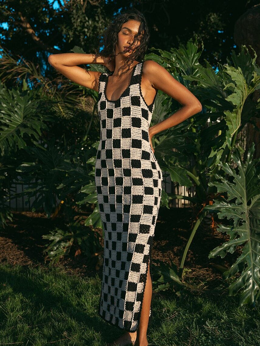 A woman stands outdoors in a checkered sleeveless dress, posing with one hand behind her head and surrounded by lush greenery.