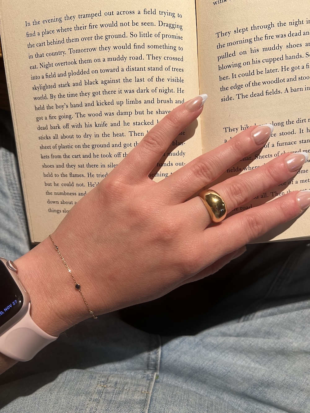 A hand wearing a gold ring and a bracelet holds open a book. A smartwatch is on the wrist.