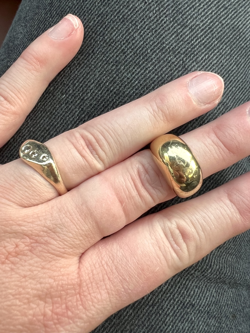 Close-up of a hand wearing two rings; one silver with small raised dots on the left hand's index finger, and one smooth, thick gold band on the middle finger, against a dark fabric backdrop.