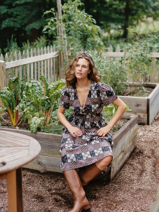 A person in a patterned dress and brown boots is sitting on the edge of a raised garden bed with green plants, with a picket fence and trees in the background.