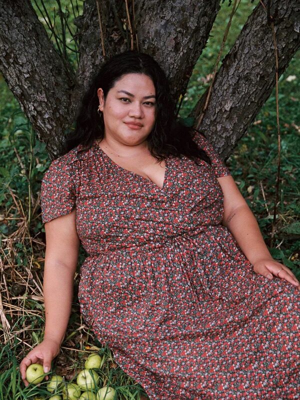 A woman in a floral dress sits against a tree in a grassy area, holding some green apples in her hand and having more apples on the ground beside her.