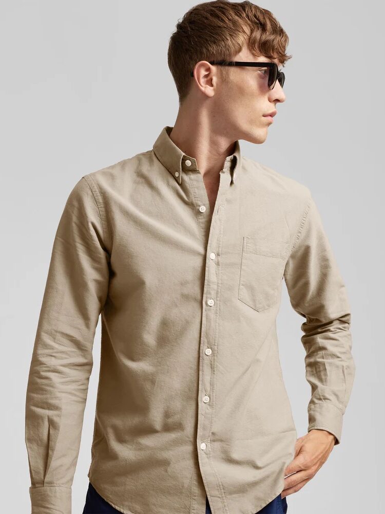 A person wearing sunglasses and a beige long-sleeve button-down shirt looks to the side against a plain background.