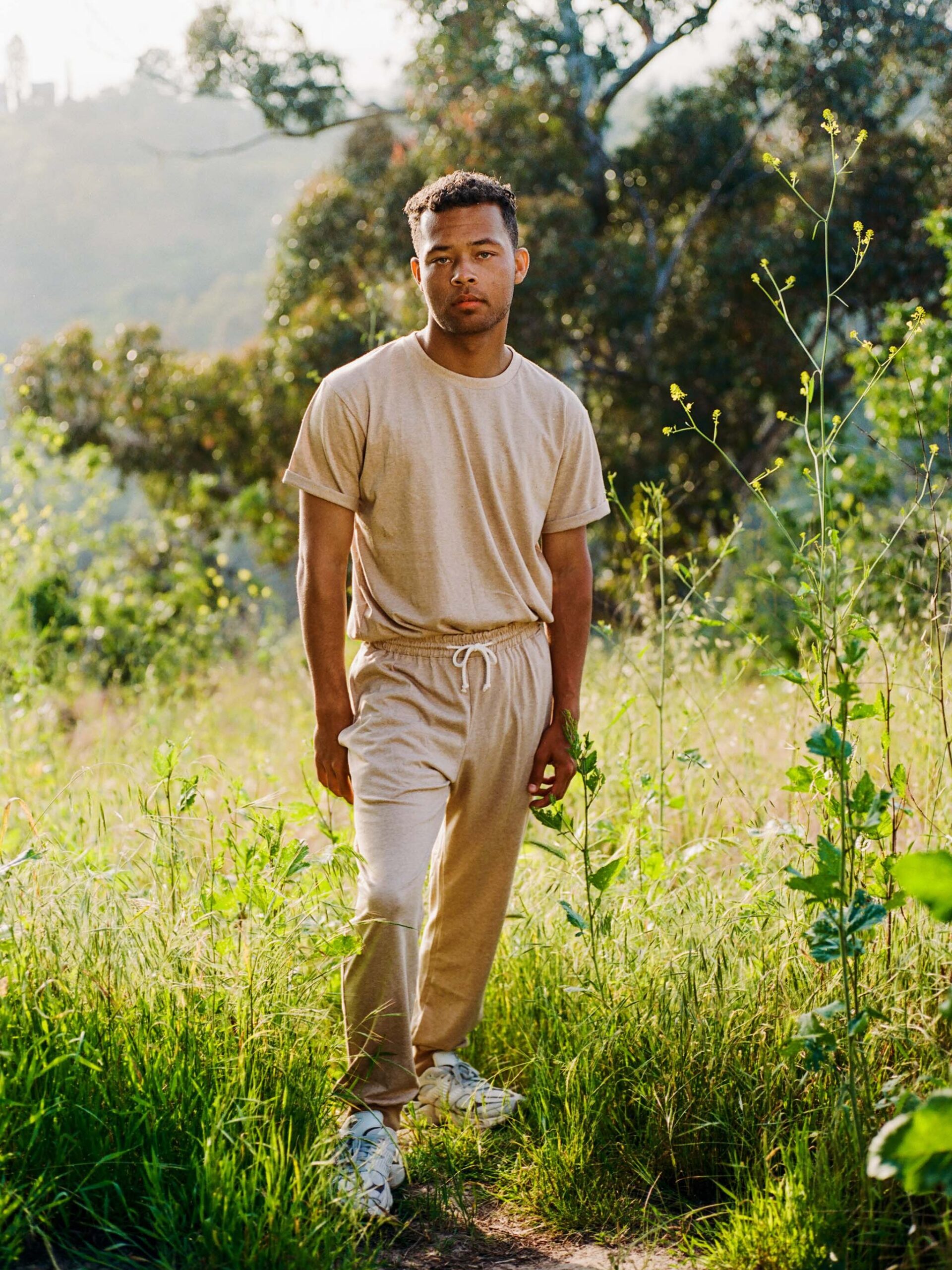 A person wearing a beige t-shirt and matching pants stands on a path in a grassy outdoor area, surrounded by greenery and trees.