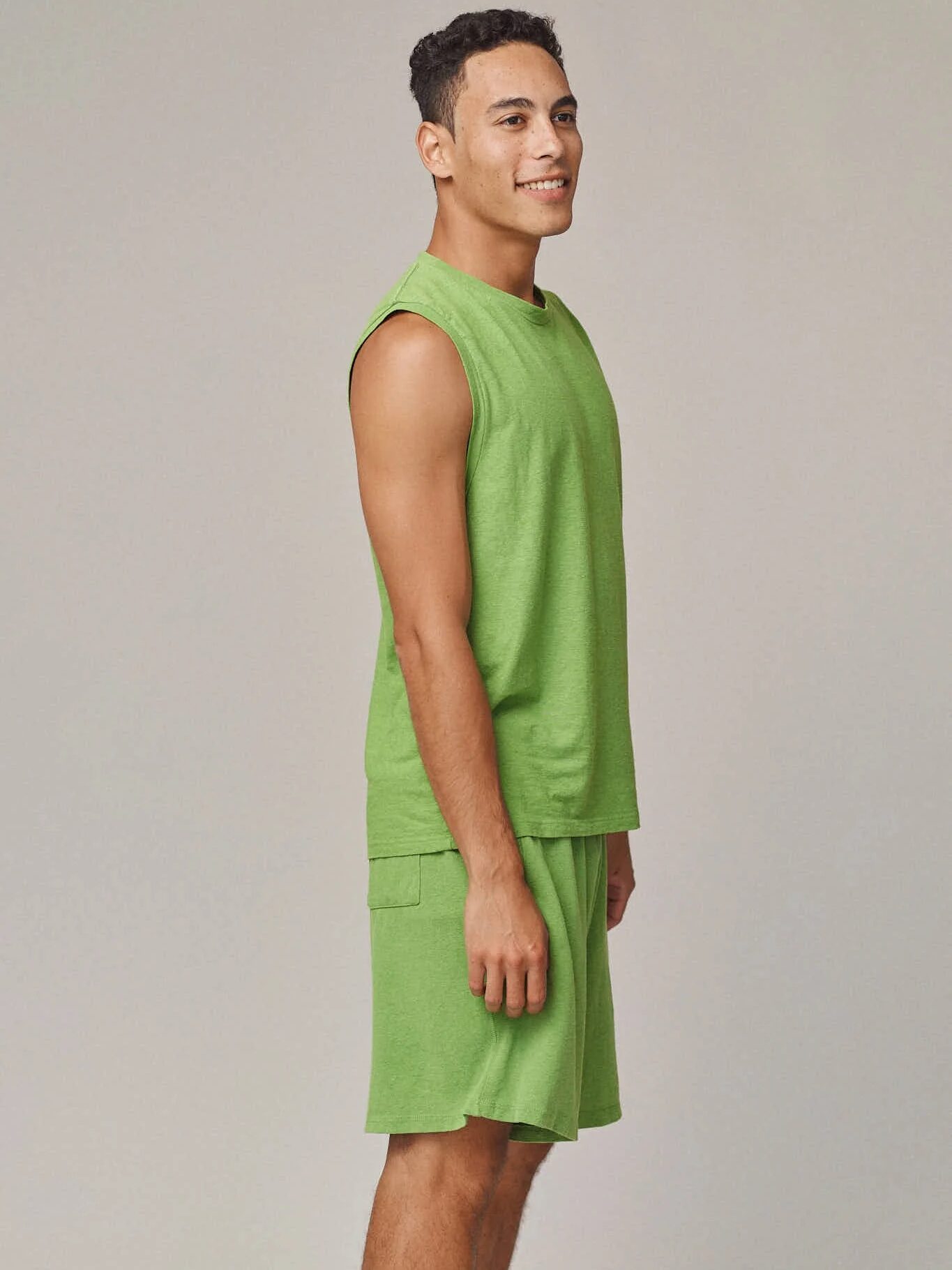 A smiling man stands in profile wearing a green sleeveless shirt and matching green shorts against a plain background.