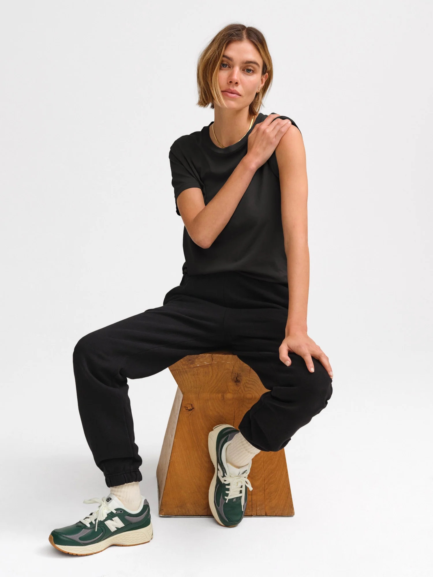 A person with short hair sits on a wooden block, wearing a black T-shirt, black pants, white socks, and green-and-white sneakers.