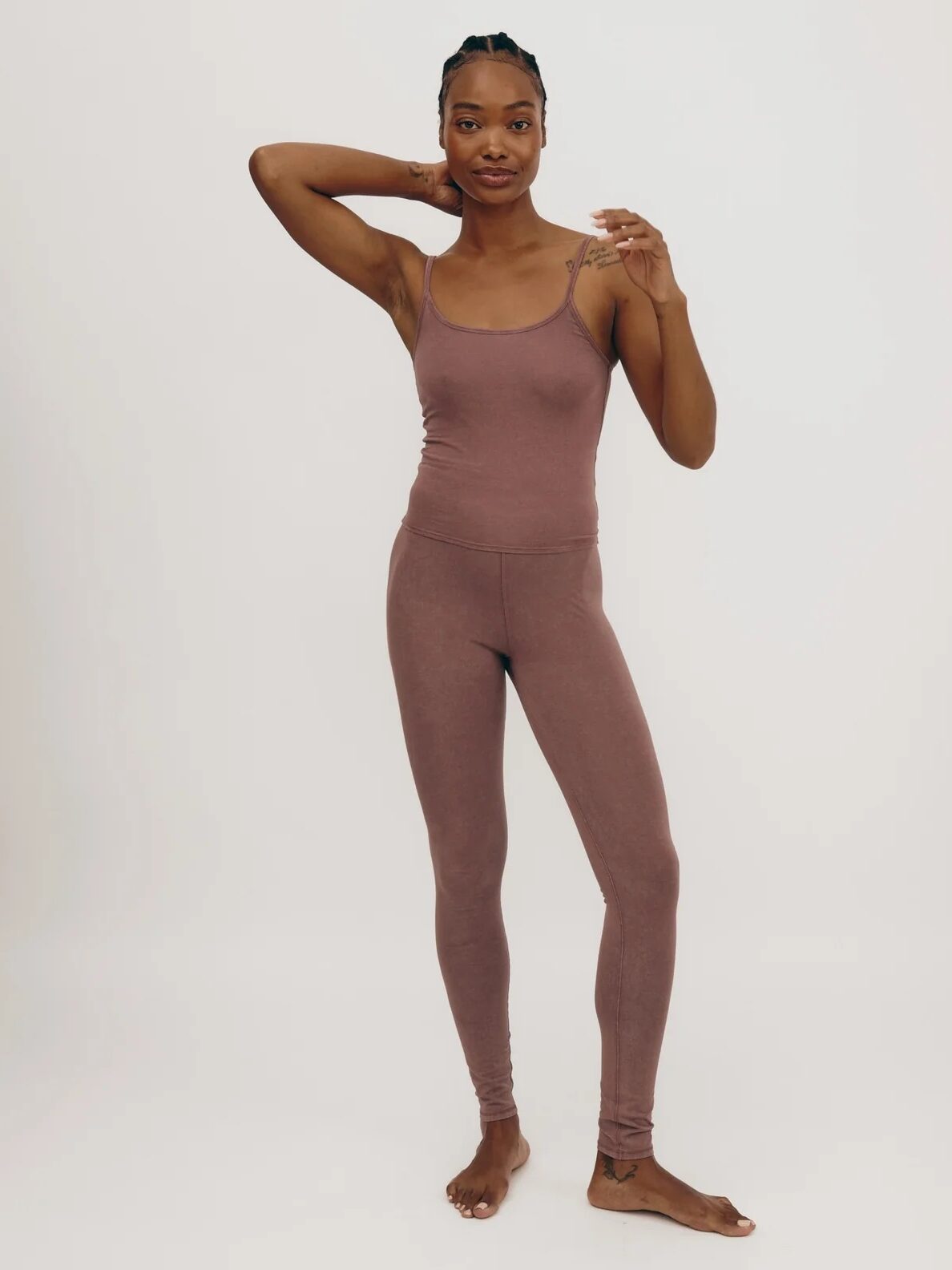 A person stands against a plain background wearing a mauve full-body athletic outfit, with one arm bent at the elbow and the other touching her neck.