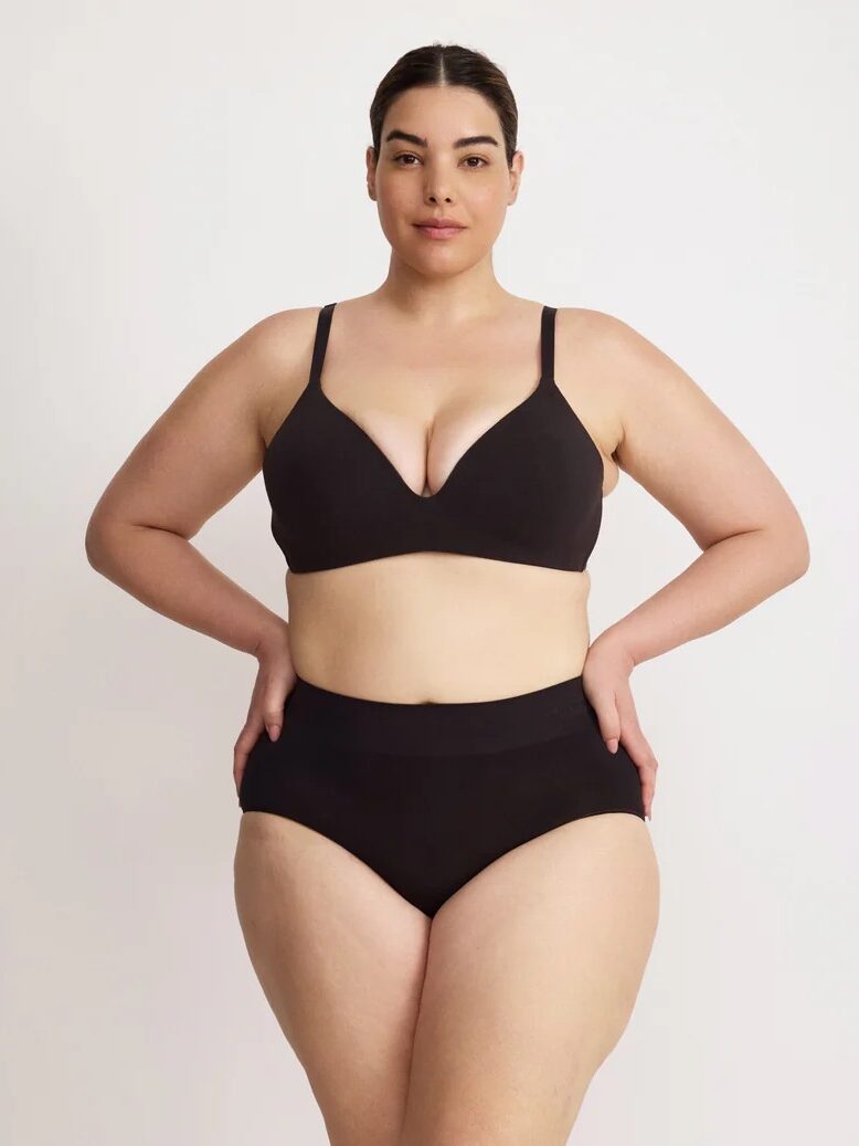 A person stands against a plain background, wearing a black bra and black high-waisted underwear, with hands on hips.