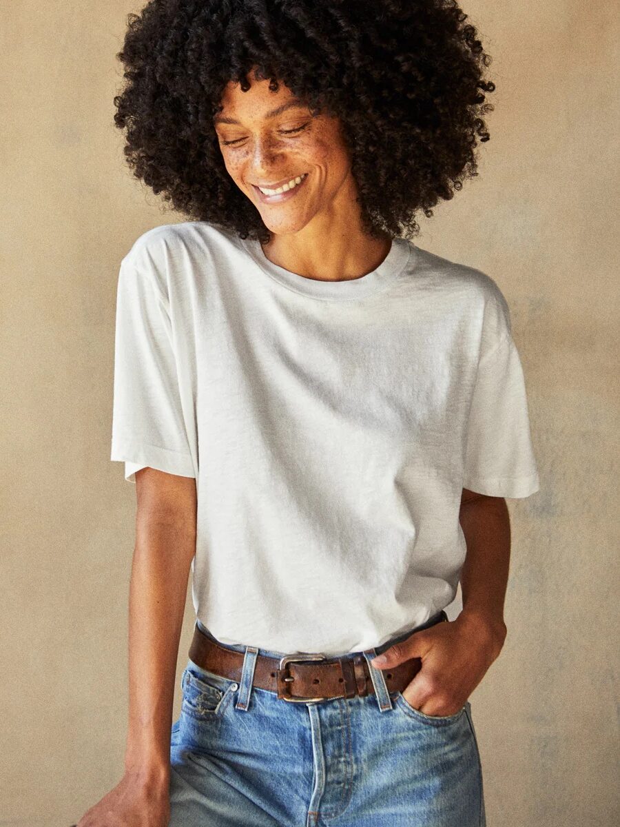 A person with curly hair is smiling, wearing a white t-shirt tucked into blue jeans with a brown belt. The background is plain and neutral.