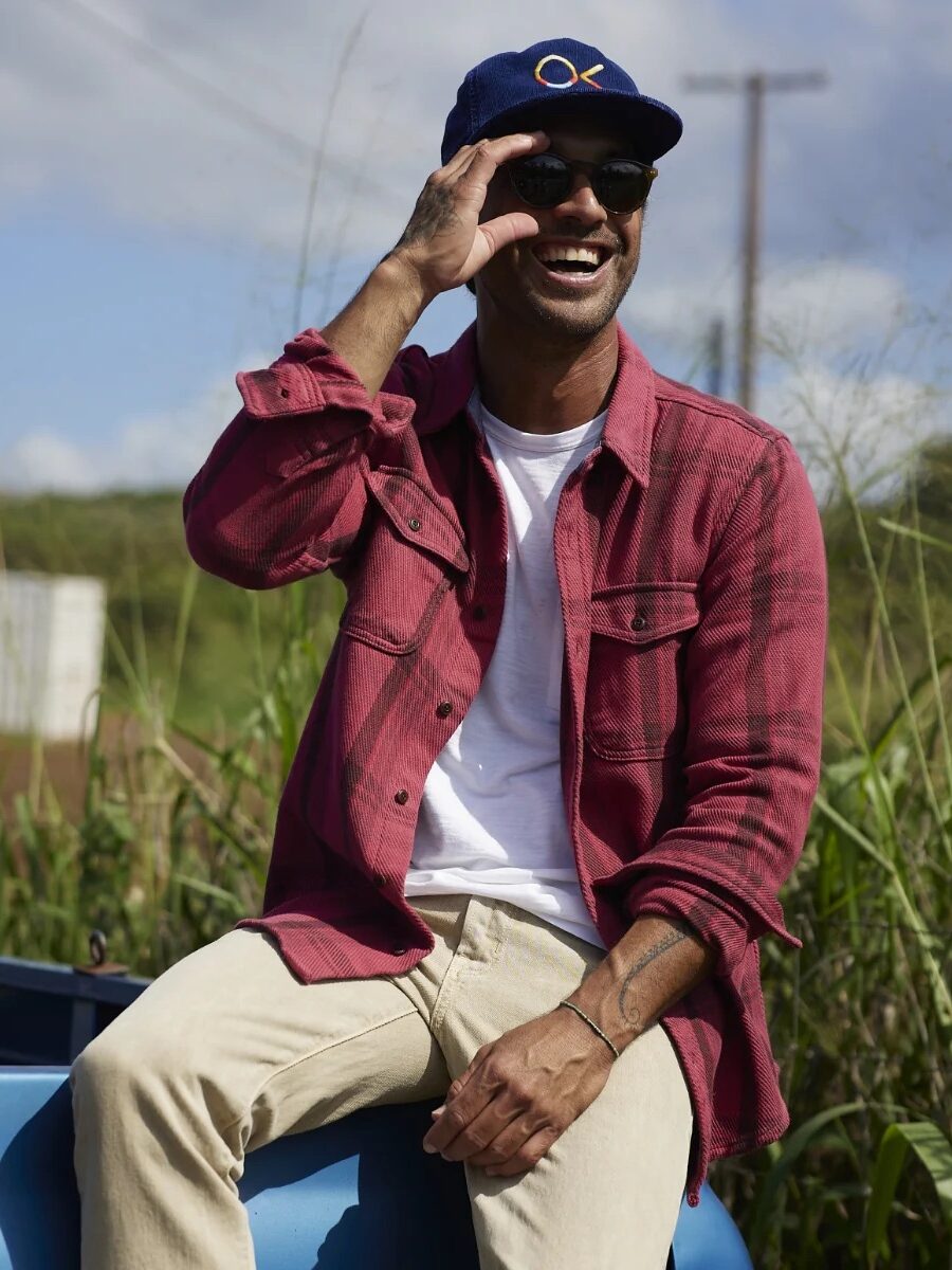 Person sitting on a blue vehicle in an outdoor setting, wearing a red shirt, beige pants, a blue cap, and sunglasses, while smiling and holding up fingers around one eye.