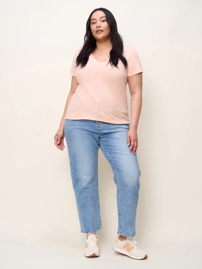 A woman stands against a plain background, wearing a light pink T-shirt, blue jeans, and white sneakers with orange details. She has long black hair and a neutral expression.