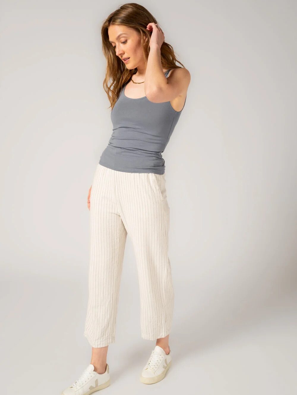 Woman wearing a grey tank top, light striped pants, and white sneakers stands in a neutral pose against a plain background.